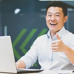 A person thumbs-upping in front of a laptop while smiling at the camera